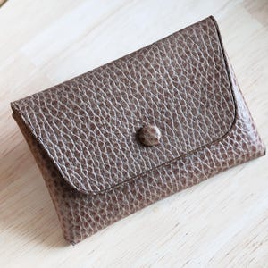 Peut inclure: Un portefeuille en cuir marron avec une surface texturée et une fermeture à bouton. Le portefeuille rectangulaire a un rabat incurvé et est posé sur une surface en bois clair.