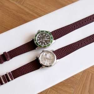 May include: Two wristwatches with brown leather bands on a white surface. The watch on the left has a green face with black markings and a silver bezel. The watch on the right has a white face with black Roman numeral markings and a silver bezel.