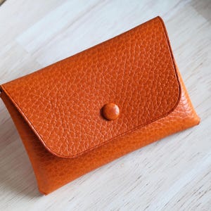Peut inclure: Un porte-cartes en cuir orange avec une surface texturée et une fermeture à bouton. Le portefeuille rectangulaire a un rabat fixé par un petit bouton rond. Le porte-cartes est posé sur une surface en bois de couleur claire.