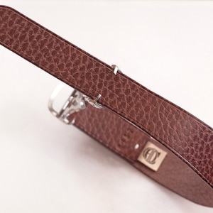 Könnte beinhalten: Ein brauner Ledergürtel mit strukturierter Oberfläche und silberfarbener Schnalle. Der Gürtel hat eine rechteckige goldfarbene Schnalle mit den Initialen "CD". Der Gürtel ist diagonal vor weißem Hintergrund angeordnet.