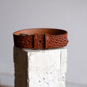 May include: A brown leather belt with a silver buckle. The belt is on a white concrete block.