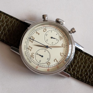 May include: A vintage-style chronograph watch with a cream-colored face, featuring black hands and numerals, and two sub-dials. The watch has a stainless steel case and a dark green leather strap. The watch face is approximately 1.5 inches in diameter.