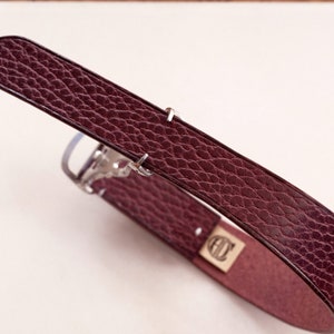 May include: A burgundy leather eyeglass case with a textured surface. The case has a rectangular shape with a flap closure and a silver-tone metal hinge. A small gold-tone metal plate with an emblem is visible.