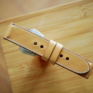 Puede incluir: Una correa de reloj de cuero marrón claro con costuras blancas. La correa tiene una hebilla y está diseñada para un reloj.