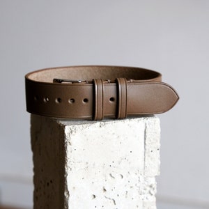 May include: A brown leather belt with a silver buckle and multiple adjustment holes. The belt is displayed on a textured, off-white surface. The leather appears to be a solid color with a matte finish.