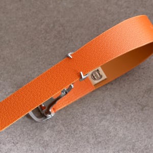 May include: An orange leather belt with a silver buckle. The belt has a textured surface and a small rectangular tag with a logo. The belt is angled across a gray surface.