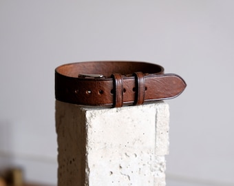 May include: A brown leather watch strap with a silver buckle. The strap is on a white concrete block.