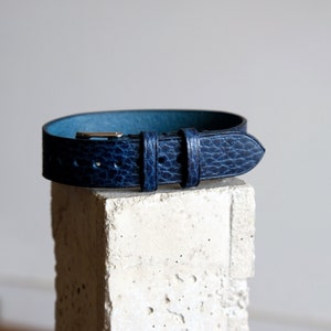 May include: A dark blue leather belt with a textured finish. The belt is shown on a light gray textured surface.