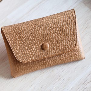 Peut inclure: Un petit portefeuille en cuir beige avec une surface texturée. Le portefeuille a une fermeture à rabat sécurisée par un seul bouton rond. Le portefeuille est rectangulaire et semble vide.