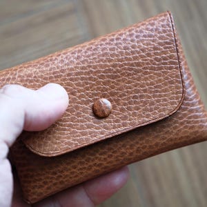 Peut inclure: Un petit portefeuille en cuir marron avec une surface texturée. Le portefeuille a une fermeture à rabat sécurisée par un seul bouton rond. Le portefeuille est tenu dans une main.