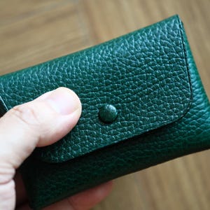 Peut inclure: Un portefeuille en cuir vert foncé avec une surface texturée. Le portefeuille a une fermeture à rabat sécurisée par un bouton assorti. Le portefeuille est tenu dans une main, mettant en valeur son design compact. Le cuir semble être de haute qualité.