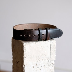 May include: A brown leather belt with a black leather tip and a silver buckle. The belt is on a white concrete block.