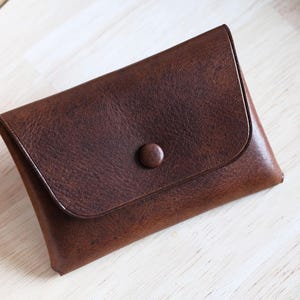 Peut inclure: Un portefeuille en cuir marron foncé avec fermeture à rabat et bouton. Le portefeuille rectangulaire présente une surface texturée. Il est posé sur une surface en bois clair. Idéal pour cartes ou argent.