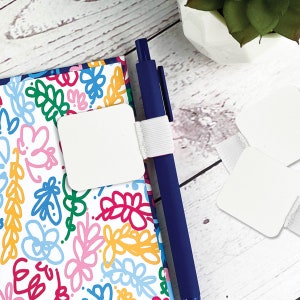 May include: A white notebook with a colorful floral pattern and a white pen holder with a white elastic band. The pen holder is attached to the notebook and holds a blue pen. There are three white square sticky notes on a white wooden surface.