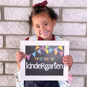 May include: A young girl with a burgundy bow in her hair holds a white framed chalkboard with colorful pennants and the text "FIRST DAY OF kindergarten".