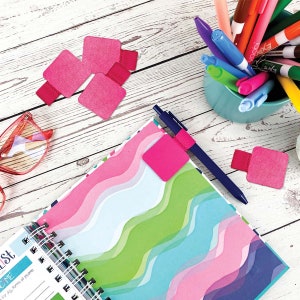 May include: Pink felt pen holders with a blue pen and a notebook with a colorful wavy pattern. The notebook has a silver spiral binding and the words "To-Do List" and "Income" are visible on the page.