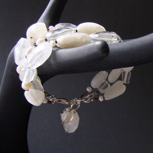 May include: A white gemstone bracelet with a silver clasp and a small white charm. The bracelet is made of various shapes and sizes of white stones.