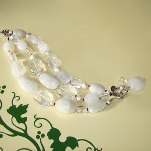 May include: A three-strand necklace made of white and clear gemstone beads. The necklace features a mix of oval and rectangular shaped beads.