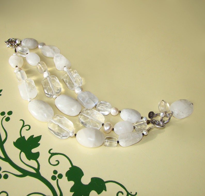 May include: A three-strand necklace made of white and clear gemstone beads. The necklace features a mix of oval and rectangular shaped beads.