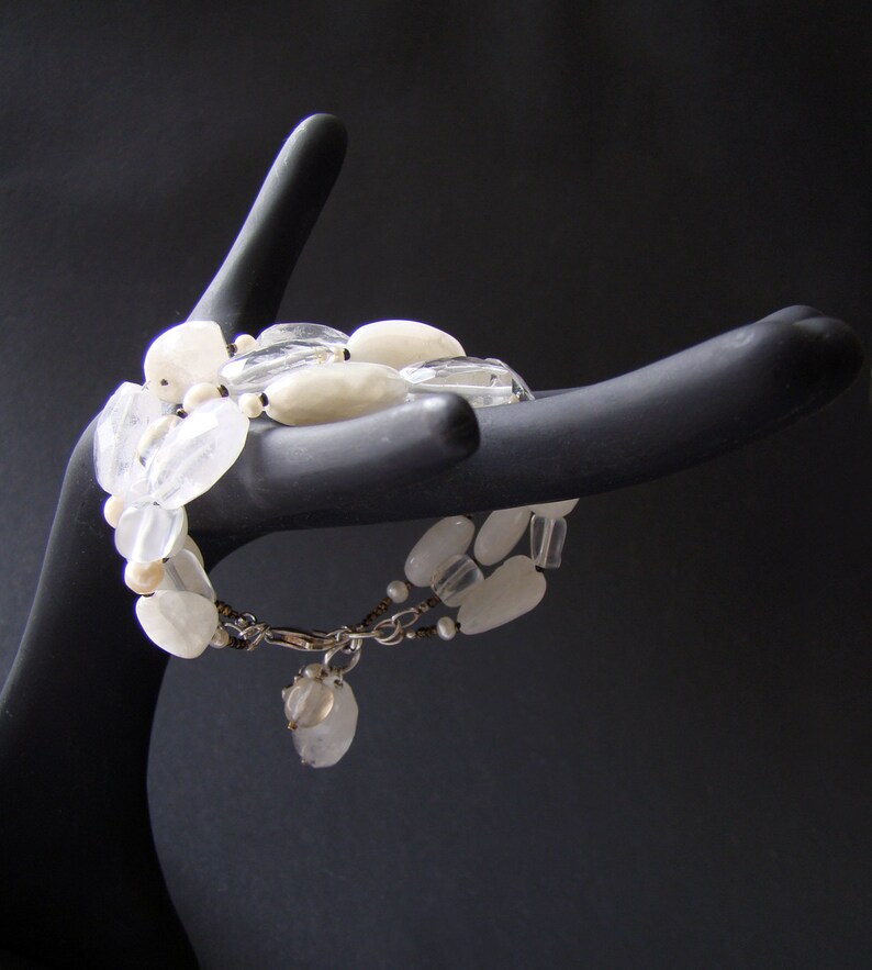 May include: A white gemstone bracelet with a silver clasp and a small white charm. The bracelet is made of various shapes and sizes of white stones.