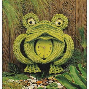 May include: A green crocheted frog with large eyes and a clock face in its belly. The frog is on a background of brown wood and green plants. The text "Rudi Ribbet" and "the friendly frog" is above the frog. The text "from the O. Henry World of Craft Ideas" is below the frog.