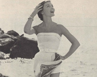Pode incluir: Uma mulher em um maiô branco sem alças com um cinto e um sarong estampado em uma praia.