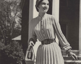 May include: A black and white photo of a woman wearing a white ribbed dress with a cardigan neckline and a matching hat. She is holding a brown handbag and standing in front of a house.