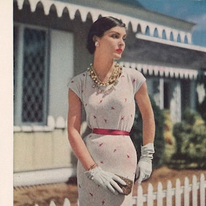 May include: A white knitted dress with a low neckline and red embroidered details. The dress is fitted and has a skirt that falls to the knees. The model is wearing white gloves and a red belt. The dress is photographed in front of a white picket fence.
