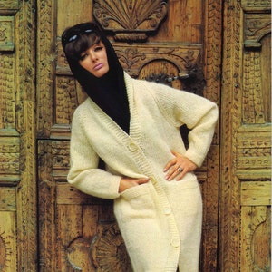 Puede incluir: Una mujer lleva un cárdigan de punto blanco con una bufanda negra y gafas de sol. El cárdigan tiene cuello en V y botones en la parte delantera. La mujer está de pie frente a una puerta de madera con tallas intrincadas.