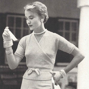 Puede incluir: Una mujer con un suéter rosa claro de envoltura con una falda a juego y un bolso blanco. Lleva un collar de perlas y pendientes.