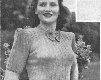 May include: A woman wearing a knitted sweater with a bow at the neckline. The sweater has a ribbed texture and a short sleeve. The woman is standing in front of a leafy background.