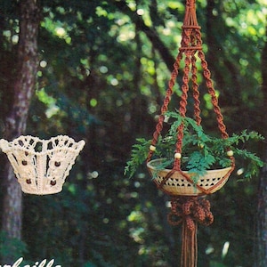 Può includere: Due portavasi in macramè, uno bianco e uno marrone, appesi ai rami degli alberi in un ambiente boschivo. Il portavasi bianco è etichettato "La Corbeille" e il portavasi marrone è etichettato "Mignon".