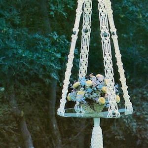 May include: A white macrame plant hanger with a round glass shelf and a tassel. The hanger is hanging from a tree branch and has a flower pot with flowers on the shelf. The text "Le Prince" is visible in the bottom right corner of the image.