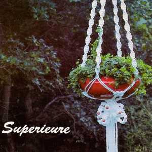 May include: White macrame plant hanger with red beads, holding a red pot with green plants. The hanger is suspended from a hook and has a long tassel at the bottom. The text "Superieure" is visible in the lower left corner of the image.
