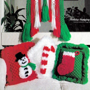 Puede incluir: Un tapiz de macramé de color verde, rojo y blanco con un tema navideño. Tres almohadas de ganchillo con diseños navideños: un muñeco de nieve, un bastón de caramelo y una media.