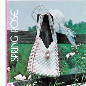 Peut inclure: Un sac à main en macramé blanc et rose avec une sangle assortie est suspendu à une clôture en bois. Un chapeau de soleil blanc avec un ruban rose repose sur la clôture. Les mots "SPRING ROSE" sont imprimés verticalement.