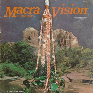 Op de afbeelding: Vintage magazinecover met een macramé plantenbak met een geweven mand met groene planten. De hanger is oranje en wit, met houten kralen. De achtergrond toont een schilderachtig uitzicht op een rivier en bergen. Tekst luidt "Macra Vision".