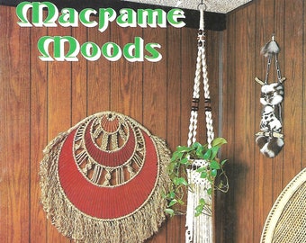 May include: A book cover with the title "Macrame Moods" in green letters. The cover features a brown wooden wall with a macrame wall hanging, a macrame plant hanger, and two potted ferns. The text on the cover also reads "Exciting New Designs Including Macra-Weaving and Macra-Sculpture".