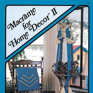 May include: A blue macrame plant hanger with white accents and a matching pillow on a wooden rocking chair. The image is from a book titled "Macrame for Home Decor II". The plant hanger has a pot of green plants inside.