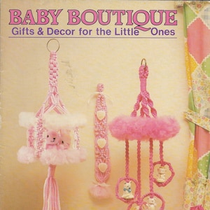 Könnte beinhalten: Ein rosa-weißes gehäkeltes Baby-Mobile mit drei hängenden weißen Hasenfiguren und einem rosa-weißen gehäkelten Kinderbett mit einer weißen Hasenfigur. Das Mobile hängt an einer rosa Decke. Das Bild zeigt auch eine rosa-weiße gehäkelte Wanddekoration mit Herzen. Der Text "BABY BOUTIQUE Gifts & Decor for the Little Ones" befindet sich oben im Bild. Der Text "MM 481" befindet sich unten im Bild.