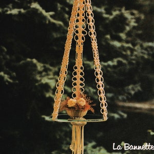 May include: A macrame plant hanger with a round glass shelf and a small bouquet of dried flowers. The hanger is made of natural colored cord and has a fringe at the bottom. The text "La Bannete" is visible in the lower right corner of the image.