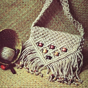May include: A beige macrame shoulder bag with a woven pattern, fringe, and wooden beads. The bag has a long strap and a triangular flap with decorative wooden beads. A basket with a ball of twine and wooden beads is next to the bag.