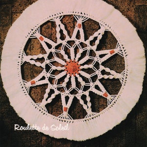 May include: A white macrame wall hanging with a circular design and red beads. The design is a sunburst pattern with a central star. The text "Roulette de Soleil" is at the bottom of the image.