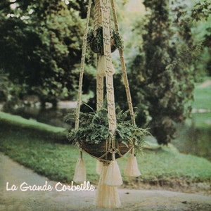 May include: A macrame plant hanger with a woven pot and tassels. The hanger is made of natural fiber rope and is hanging from a tree branch. The pot is filled with green plants. The text "La Grande Corbeille" is visible at the bottom of the image.