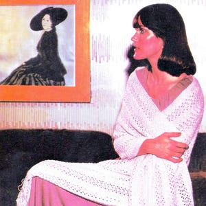 May include: A white crocheted shawl with an openwork pattern and long fringe along the edges, draped over a solid mauve-colored garment. The shawl features a repeating geometric stitch design and is worn wrapped around the upper body.