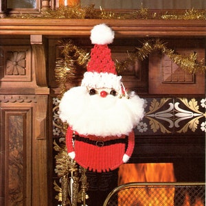 Pode incluir: Uma meia de Natal de crochê representando o Papai Noel, pendurada acima de uma lareira com fogo queimando dentro. A meia é vermelha e branca com um cinto preto e uma barba branca. A lareira é decorada com uma guirlanda dourada.