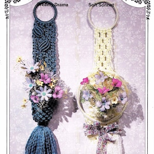 以下が含まれることがあります： 2つのマクラメの壁掛け。1つはタッセル付きの濃い青色、もう1つはクリーム色。どちらも円形のトップがあり、造花で飾られています。「Dreamy Drama」と書かれた濃い青色のものと、「Soft Sonnet」と書かれたクリーム色のものがあります。