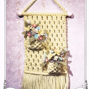 以下が含まれることがあります： 2つのポケットがあり、それぞれに造花が入ったマクラメの壁掛け。壁掛けは黄色のコードで作られており、下部にフリンジが付いています。画像の上部には「Rhapsody of Flowers」というタイトルが印刷されています。