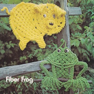 May include: A yellow crocheted pig with black eyes and a pink nose hanging on a wooden fence post. The pig is labeled "Pork Chops". Below the pig is a green crocheted frog with black eyes hanging on the same fence post. The frog is labeled "Fiber Frog".