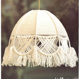 May include: A white macrame lampshade with a fringe border. The lampshade is hanging from a string. The text "La Plume Page 19" is printed below the lampshade.